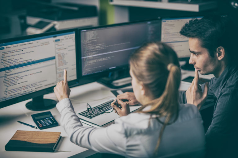 Woman and man looking a computer monitors full of scripted computer code.