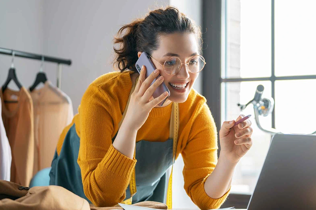 A woman in glasses looking at a laptop while holding a phone in one hand and a pen in the other